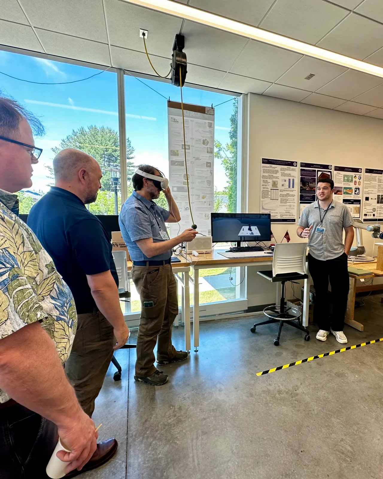A lab member wearing a VR headset demonstrates a simulation to visitors while a student presents beside a monitor